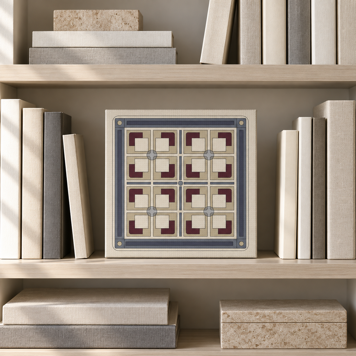 A square decorative tile or art print featuring a geometric pattern in cream, slate blue, and burgundy, displayed on a light wood bookshelf between books.