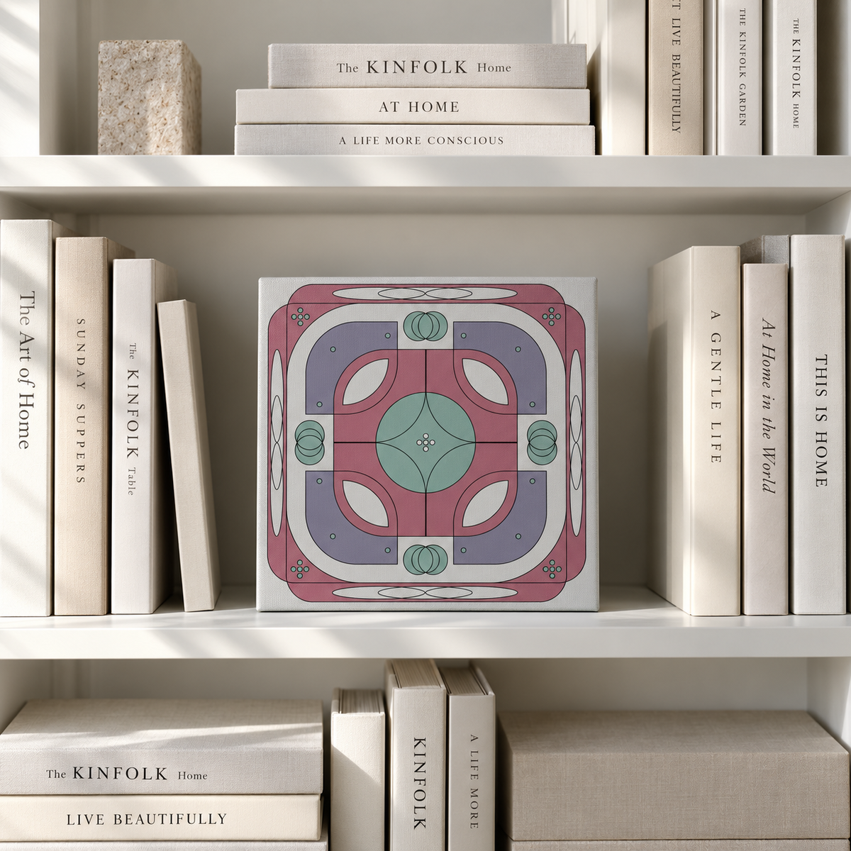 A square decorative art print or tile featuring a mid-century modern geometric pattern in dusty pink, sage green, and lavender, displayed on a white bookshelf between books.