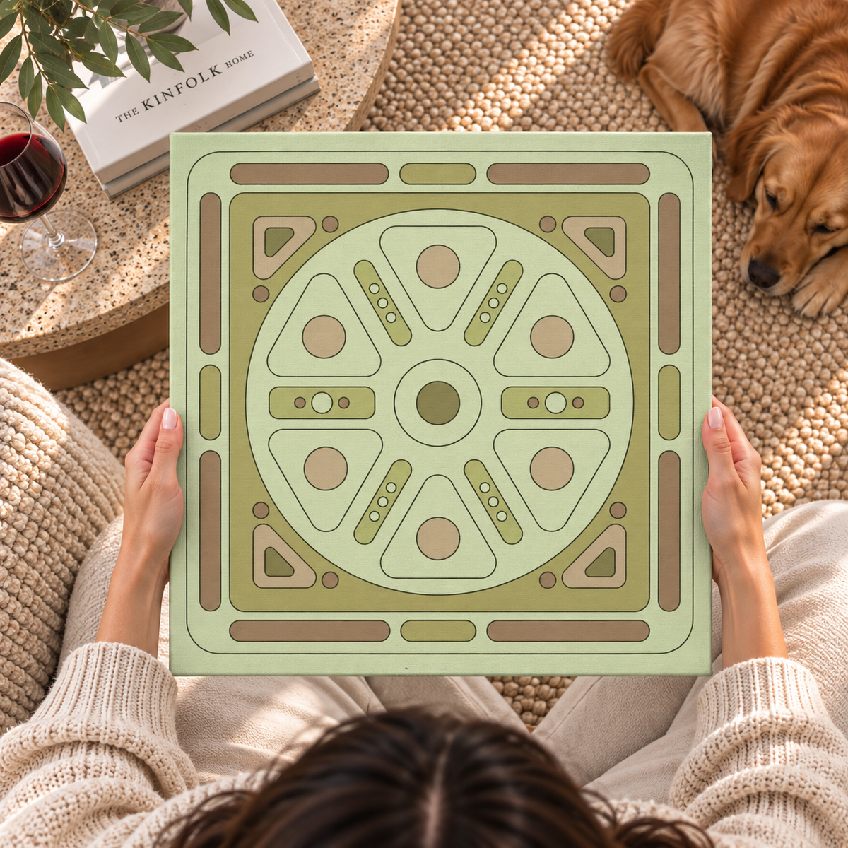 A person holds a square decorative tile or coaster featuring a retro geometric pattern in sage green, olive, and taupe. The scene is a cozy top-down lifestyle shot with a golden retriever sleeping on a woven rug, a glass of red wine, and a book in the background.