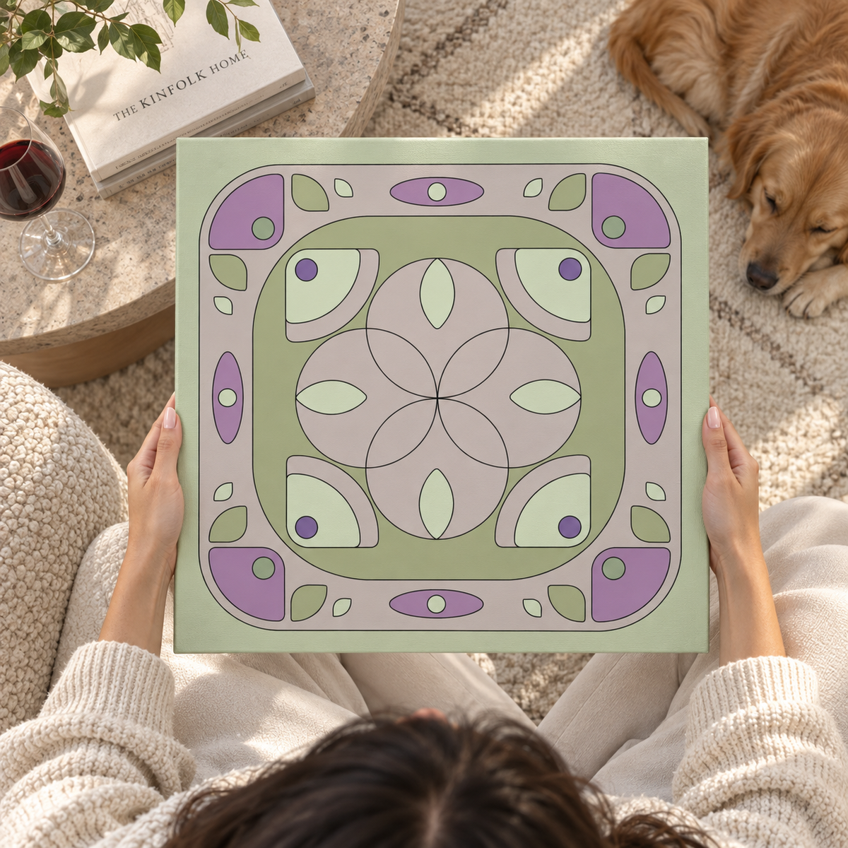 A person holds a square decorative tile or coaster featuring a retro mid-century modern geometric pattern in sage green, lavender purple, and cream. The scene is set in a cozy living room with a dog lying on a rug in the background, a glass of red wine on a side table, and a book titled 'The Knitfolk Home' nearby.